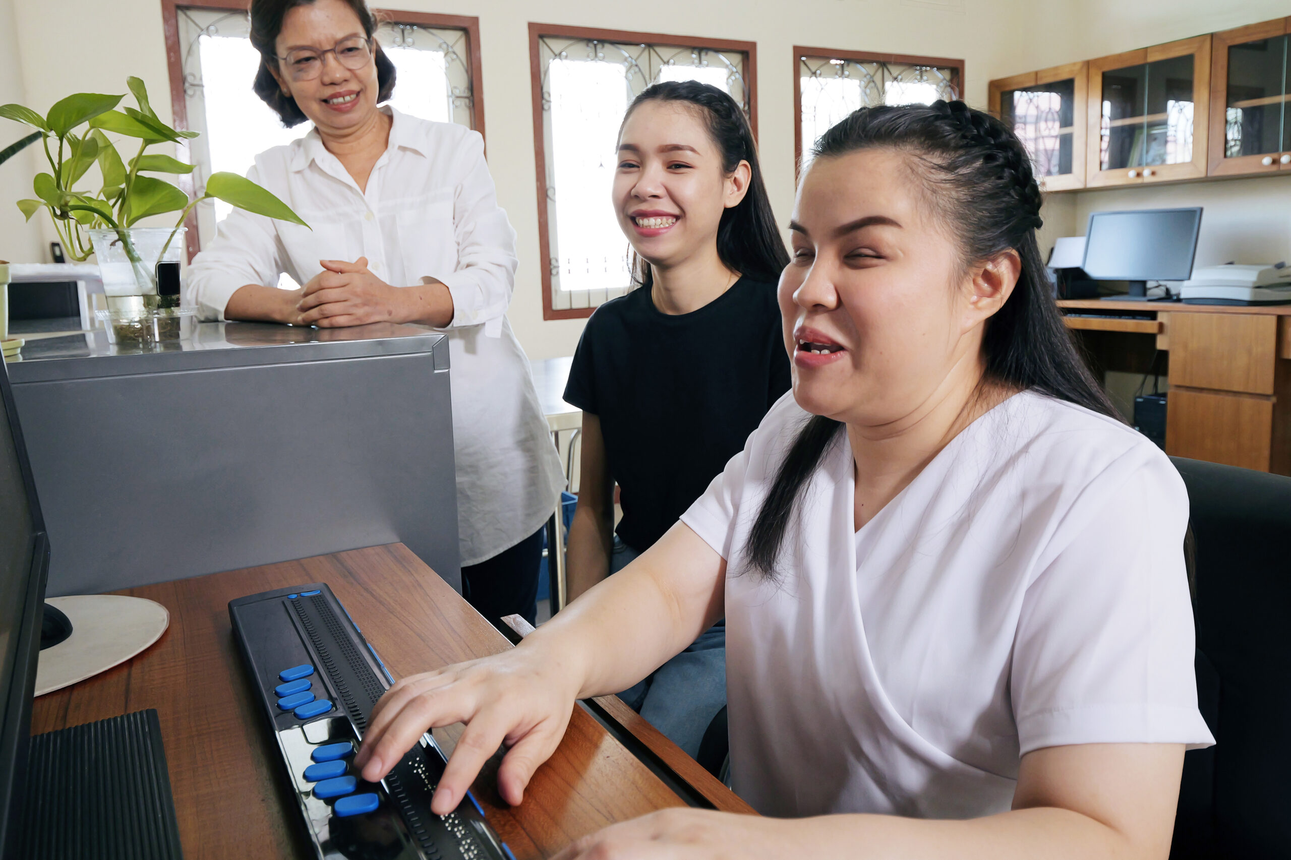 Woman at computer using accessible keyboard with two other women smiling and watching nearby